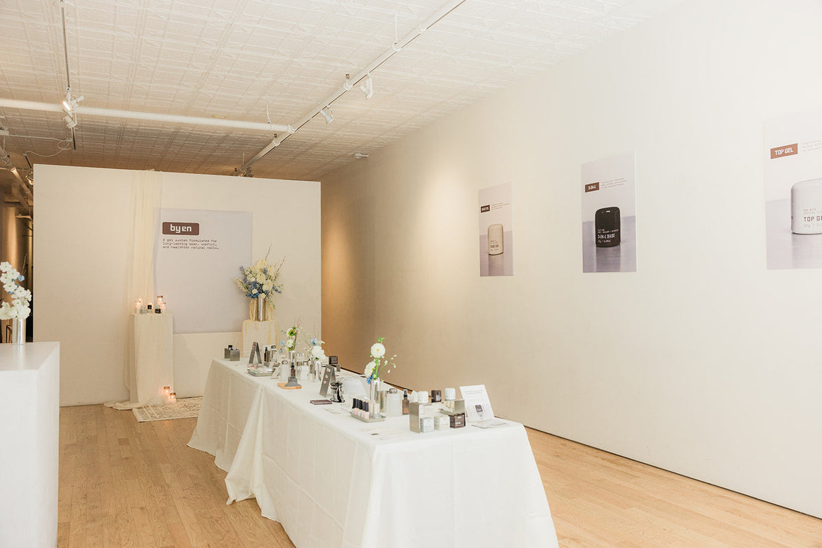 Wide view of the By En Japanese Gel Workshop, with a long table in the center of the space where nail products from Asia are displayed. There are By En posters and florals to decorate the space.